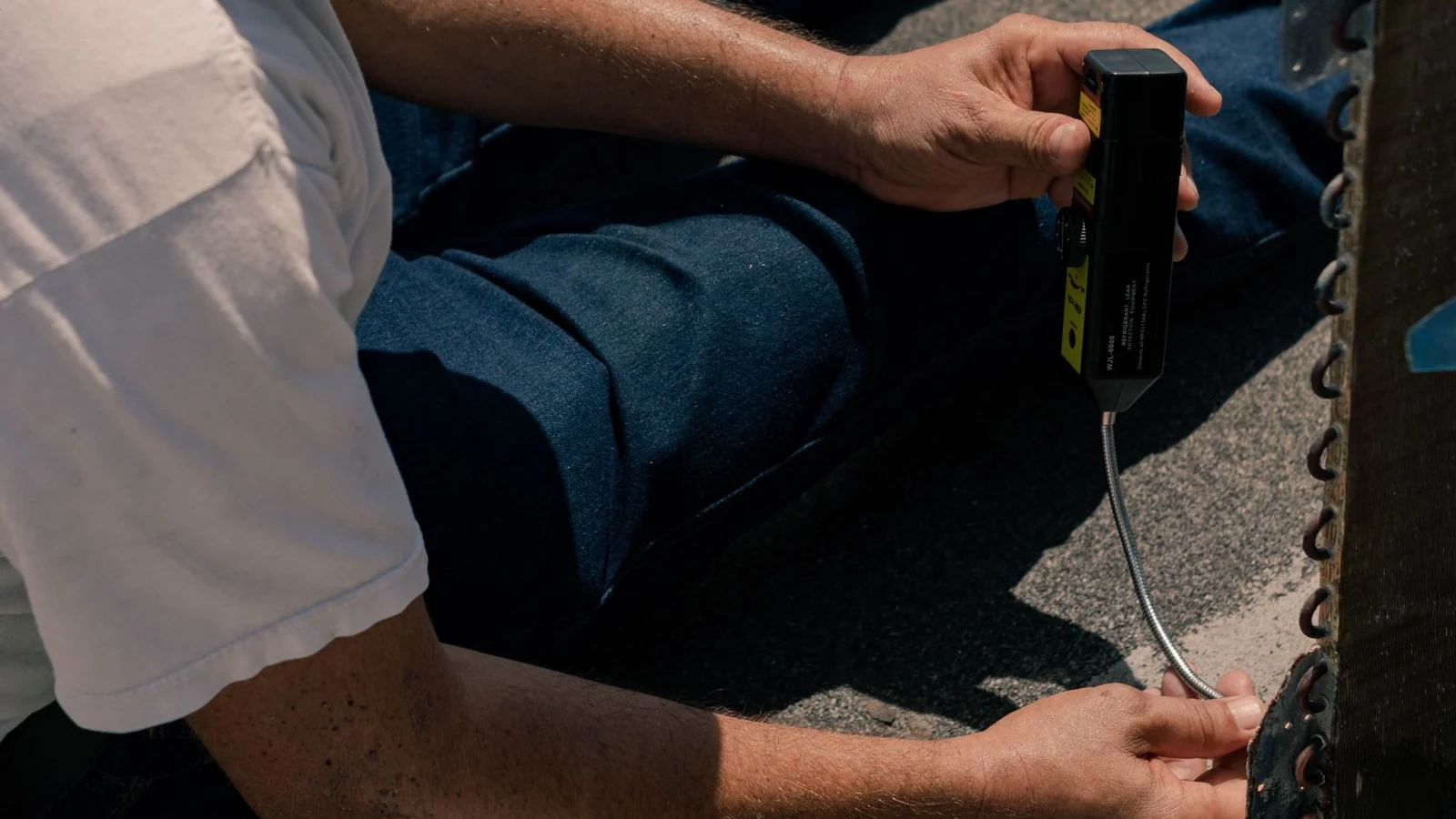 A technician sitting on a rooftop holds an electronic refrigerant leak detector against the fins of an air-conditioner coil, the technician-level check for an aircon not cold.