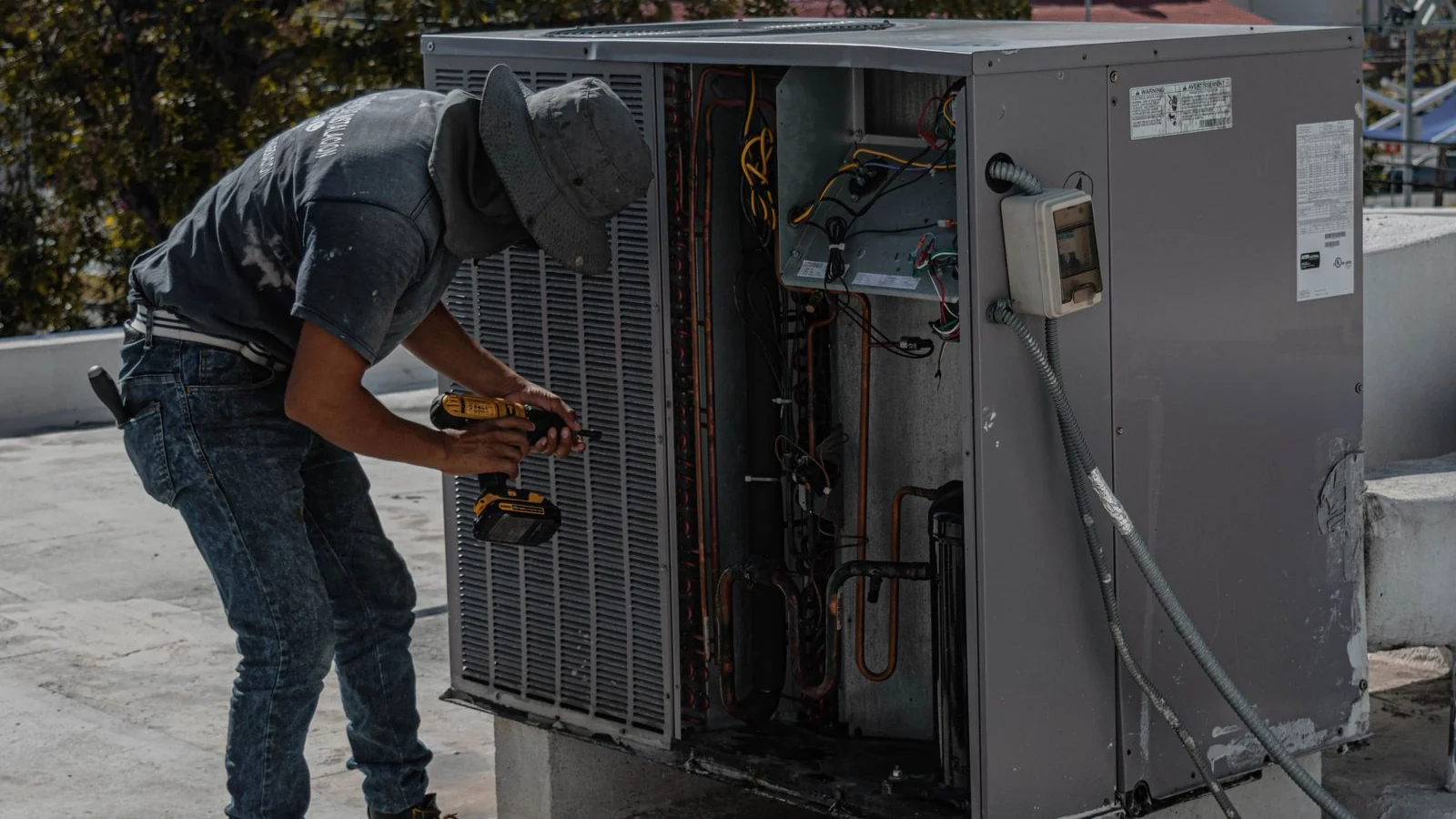 An Aircon Hero technician in a bucket hat bends over an open rooftop packaged air-conditioning unit, driving a cordless drill into its panel with trees behind him, the kind of on-site work a Daikin aircon repair call involves.