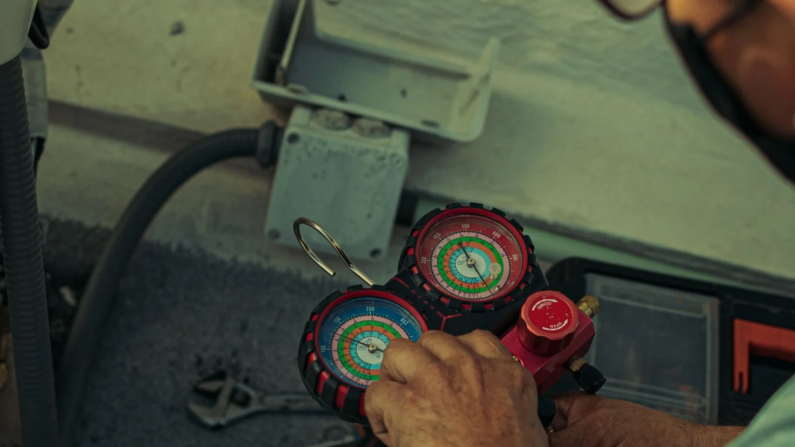 Close-up of a technician's hands on a refrigerant manifold gauge set with red and blue dials during an aircon gas top up, with a pink refrigerant cylinder and a yellow charging hose on the ground beside an outdoor unit.