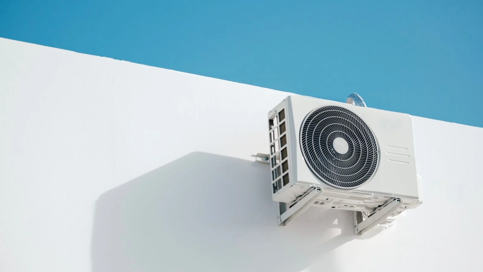 Unit kompresor aircond luar berwarna putih dipasang pada dinding putih dengan latar langit biru, jenis unit yang perlu dibersihkan semasa cuci aircond rumah.