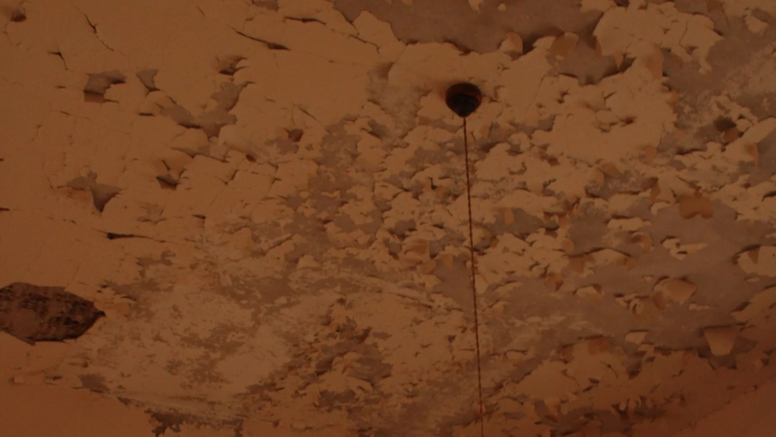 A damp brown stain spreading across a peeling ceiling in the corner of a room with a bare pendant lamp, the mark an aircon leaking water leaves overhead.