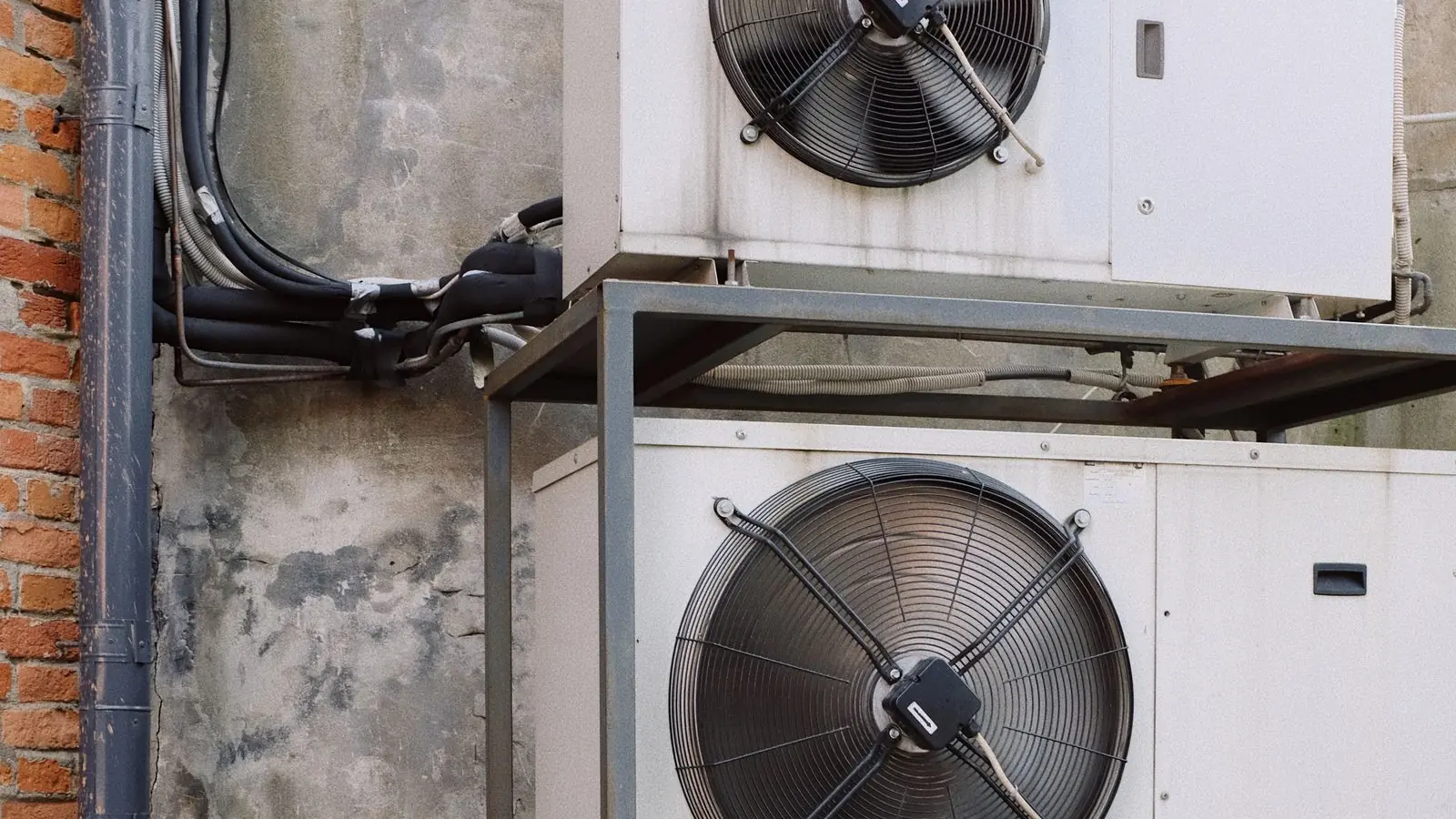 Dua unit kompresor aircond luar berwarna putih dipasang atas rangka besi di tepi dinding lama dengan paip dan pendawaian kelihatan, kerja yang dikira dalam upah pasang aircond.