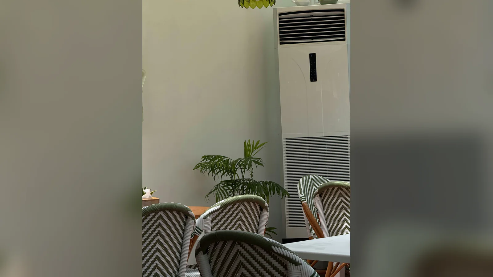 A tall white floor-standing air conditioner, the kind of unit a floor standing aircon service cleans on site, in the corner of a cafe beside a potted palm and green-and-white woven bistro chairs.
