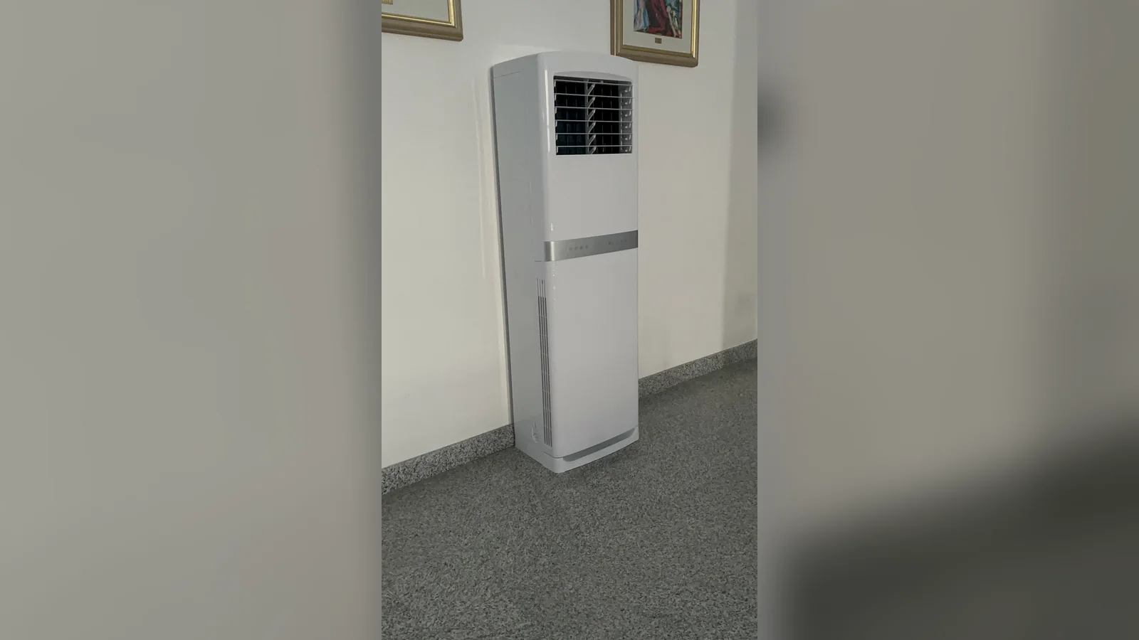 A white floor standing aircon with a slim vertical body and a silver band across the middle, standing against a white wall on a speckled grey terrazzo floor under two framed pictures.