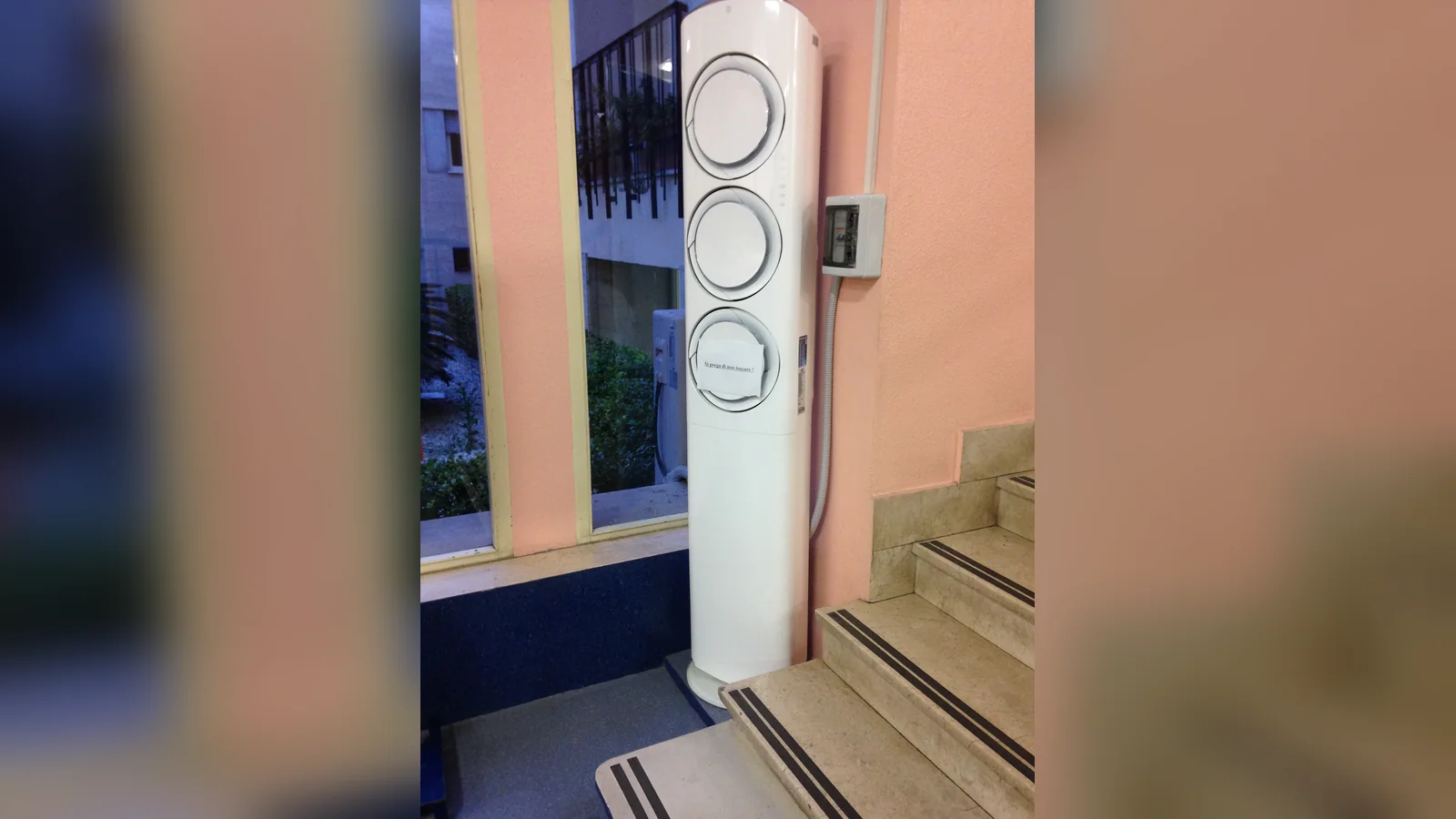 Floor standing aircon installation at the foot of a marble staircase: a tall white unit with three round air outlets beside a small wall-mounted switch box.