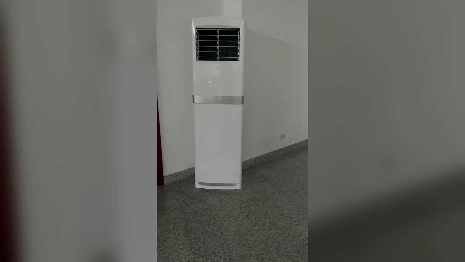 Aircond berdiri putih dengan gril di bahagian atas dan jalur perak di tengah, berdiri di sudut bilik berlantai teraso kelabu.