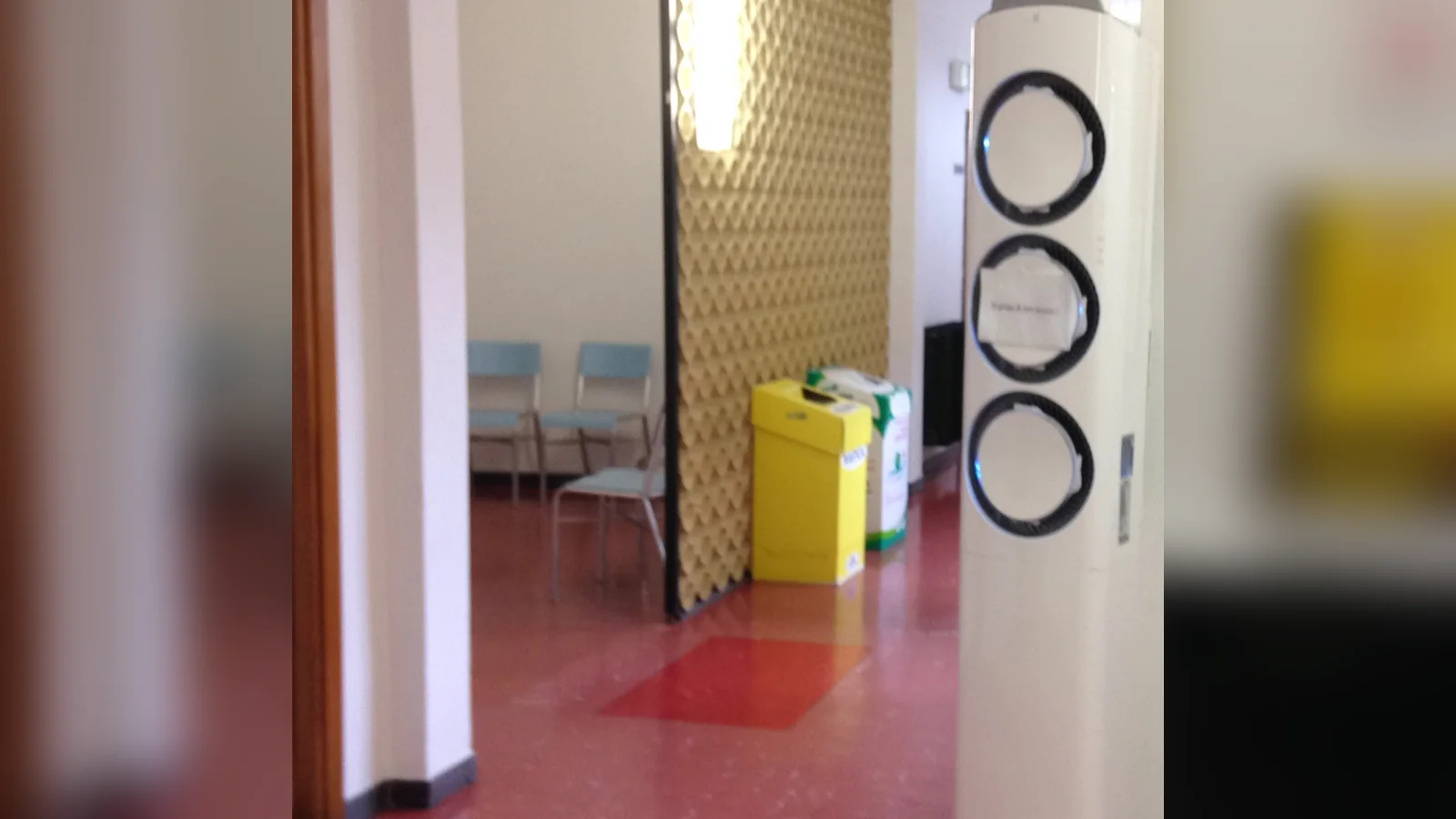 Floor standing aircon not cold? A white floor-standing air conditioner with three circular outlets stands in a public corridor with a red floor, a yellow recycling bin and waiting chairs.