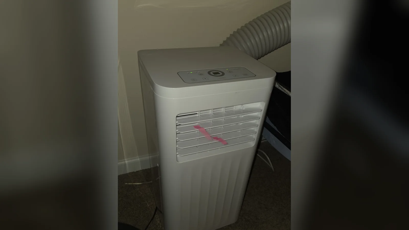 A white portable air conditioner waiting for portable aircon repair stands on a living-room carpet, its grey flexible exhaust hose rising behind it.