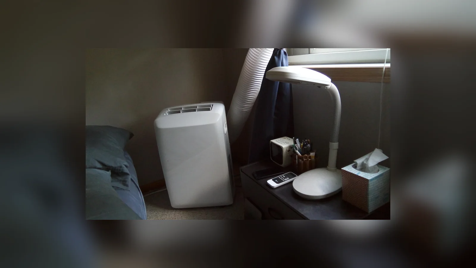 Portable aircon Malaysia bedroom setup: a compact white unit on the floor beside a bedside table and lamp, its exhaust hose running up to the window.