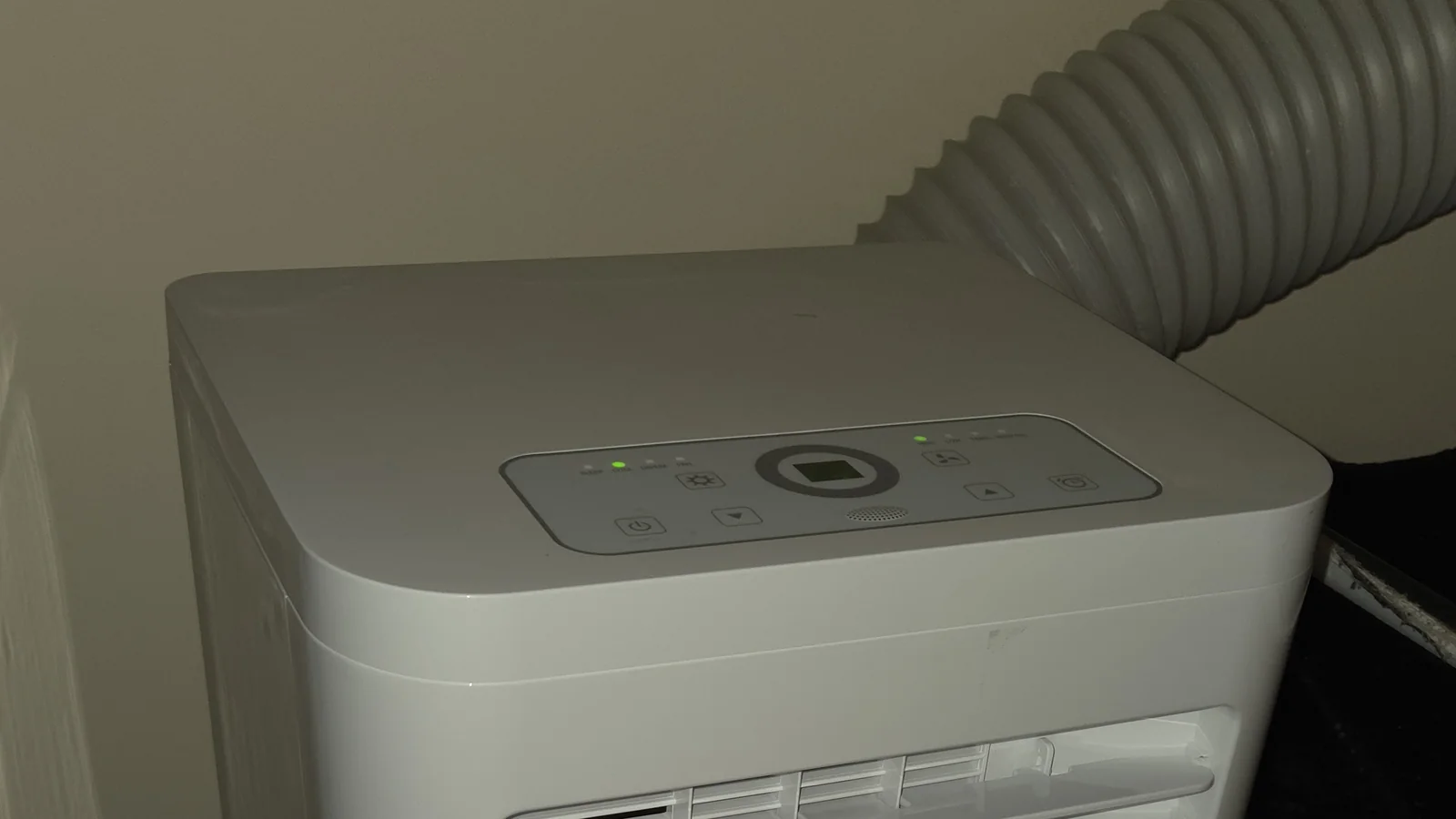 Servis portable aircond bermula di sini: panel kawalan atas dan gril hadapan sebuah unit putih, dengan hos ekzos kelabu di belakangnya.