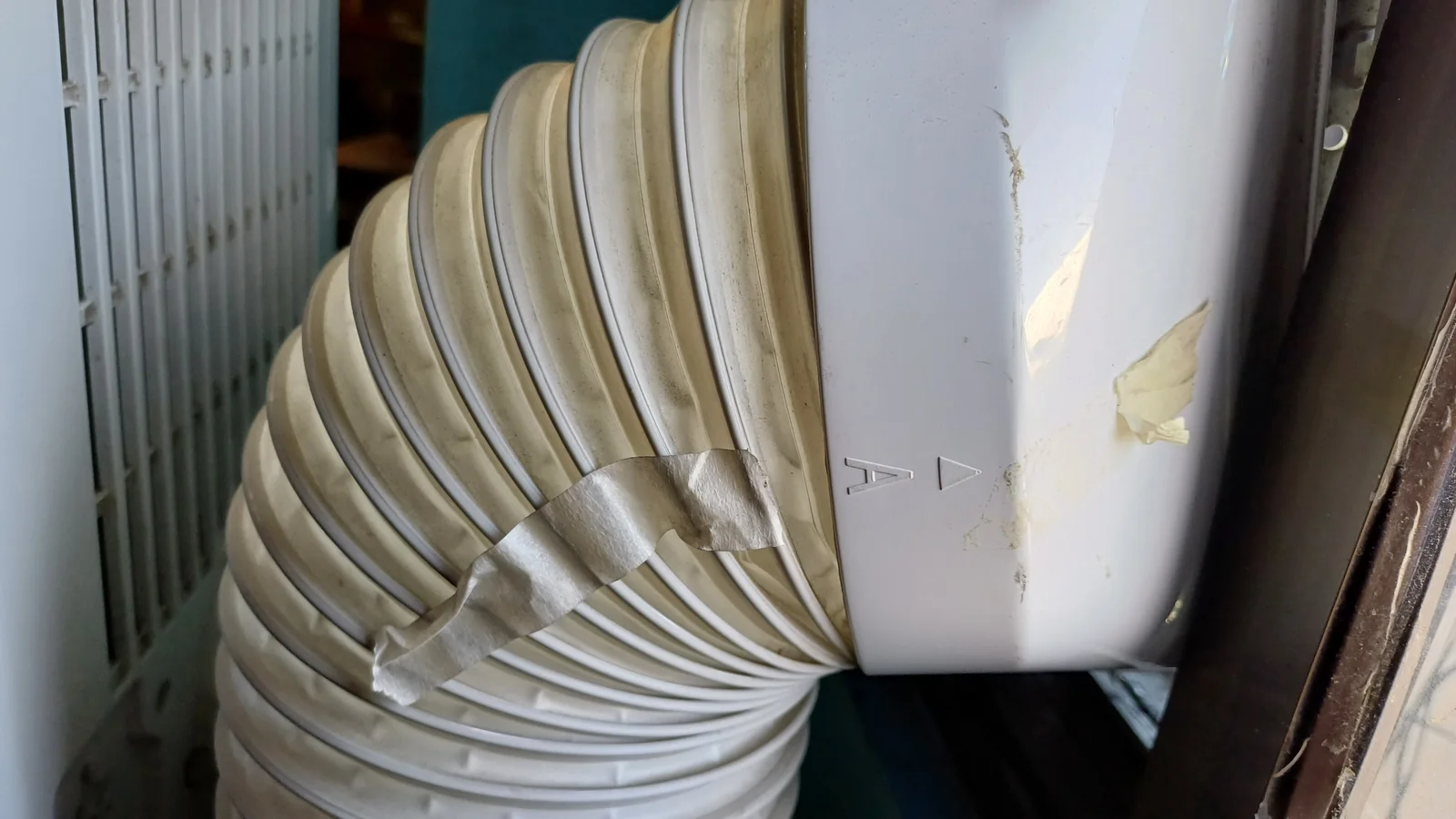 Hos ekzos putih beralun sebuah portable aircond dipasang pada tingkap dan disambung dengan pita pelekat yang sudah lusuh, salah satu tempat biasa portable aircond keluar air.