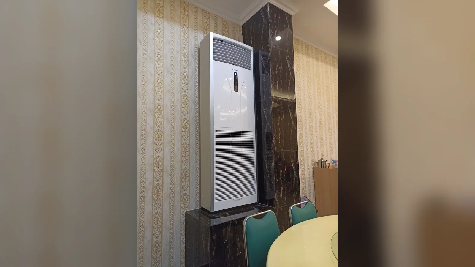 A white Daikin floor standing aircon on a black marble plinth against striped wallpaper in a dining room, with teal chairs in front.