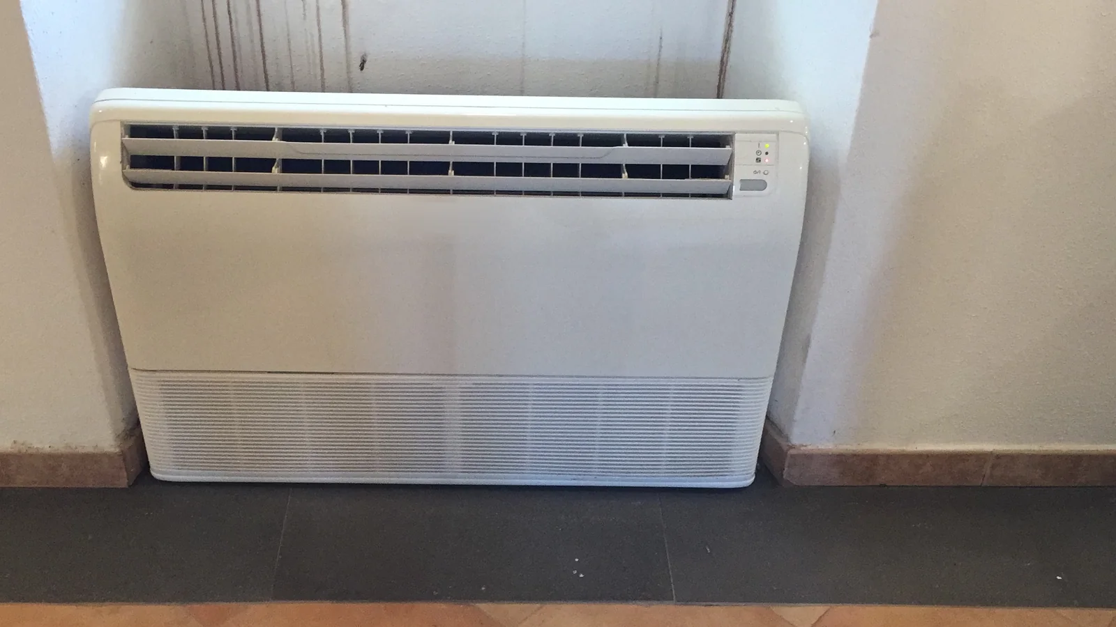 A low, knee-height white floor mounted aircon in a wall recess, with air louvres along the top and an intake grille along the base.