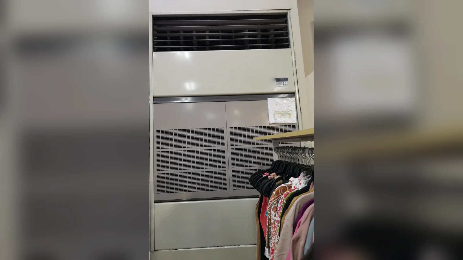 A large beige floor-standing air conditioner with a slatted top outlet and grey intake panels, standing in a clothing shop beside a rack of garments.