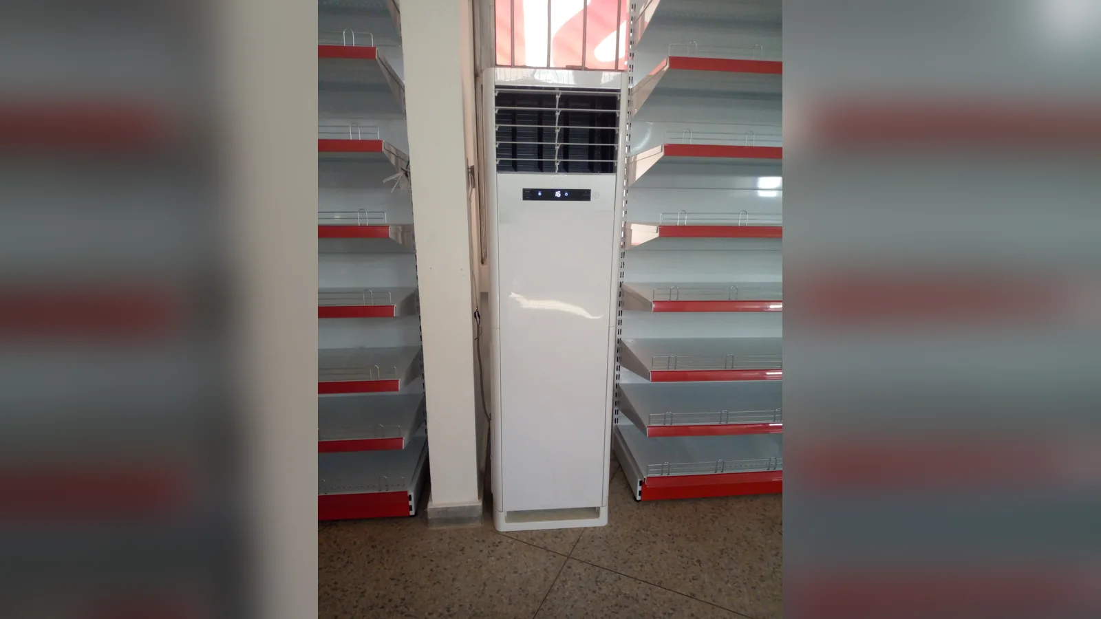 A tall white floor-standing air conditioner with a small display, standing between empty red-edged shelves on a terrazzo floor.