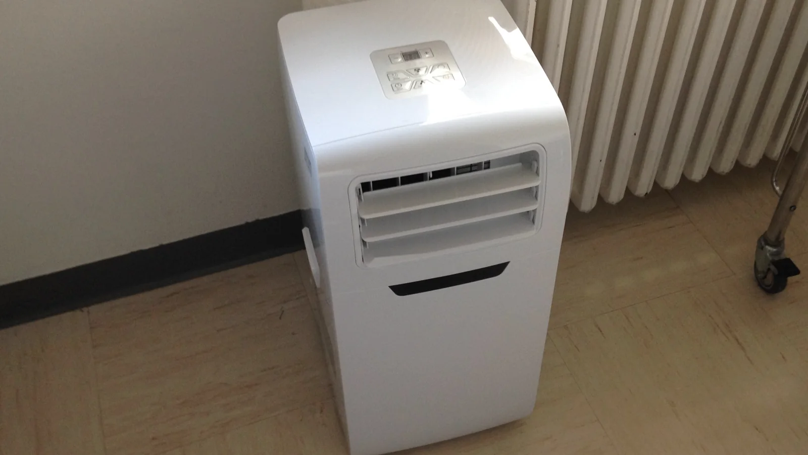 Portable aircond putih dengan panel kawalan di atas dan bilah udara di hadapan, berdiri di atas lantai jubin di sudut bilik.