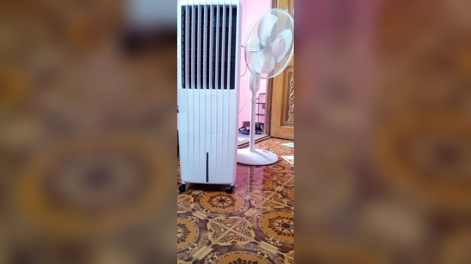 A white tower evaporative air cooler on castors beside a pedestal fan on a patterned tiled floor, the kind of unit often sold as a portable aircon without hose.