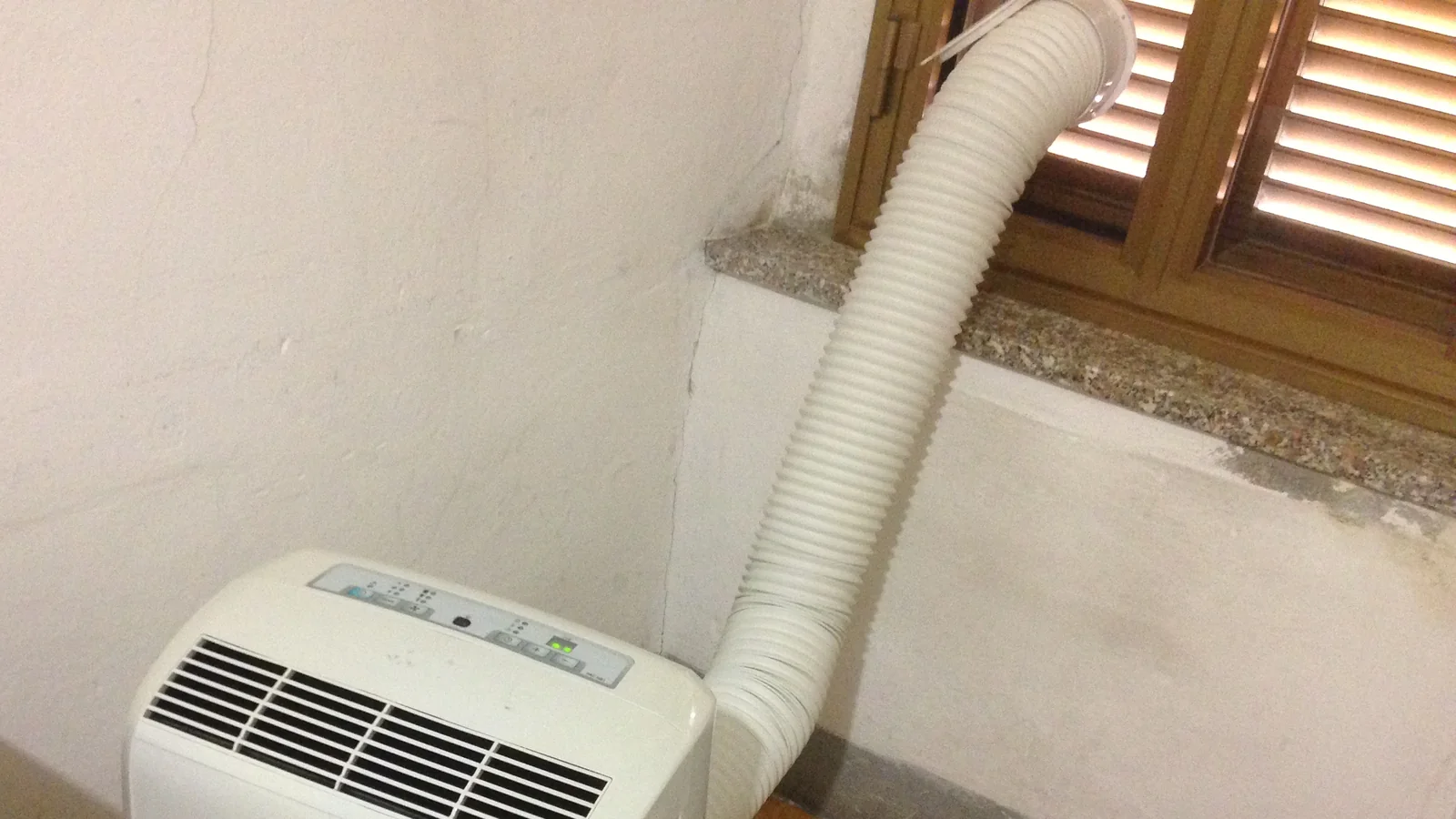 A white ribbed portable aircon hose running from the top of the unit up to a round vent in a half-open shuttered window.