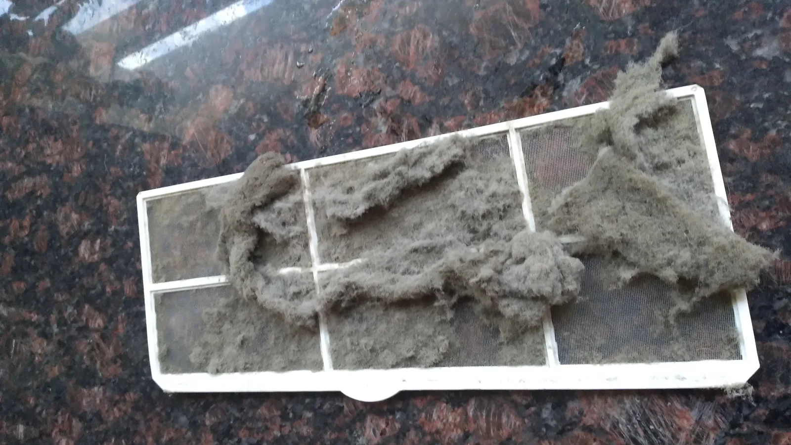 A white aircon filter clogged with thick grey dust and lint on a dark granite counter, the part every portable aircon maintenance routine starts with.
