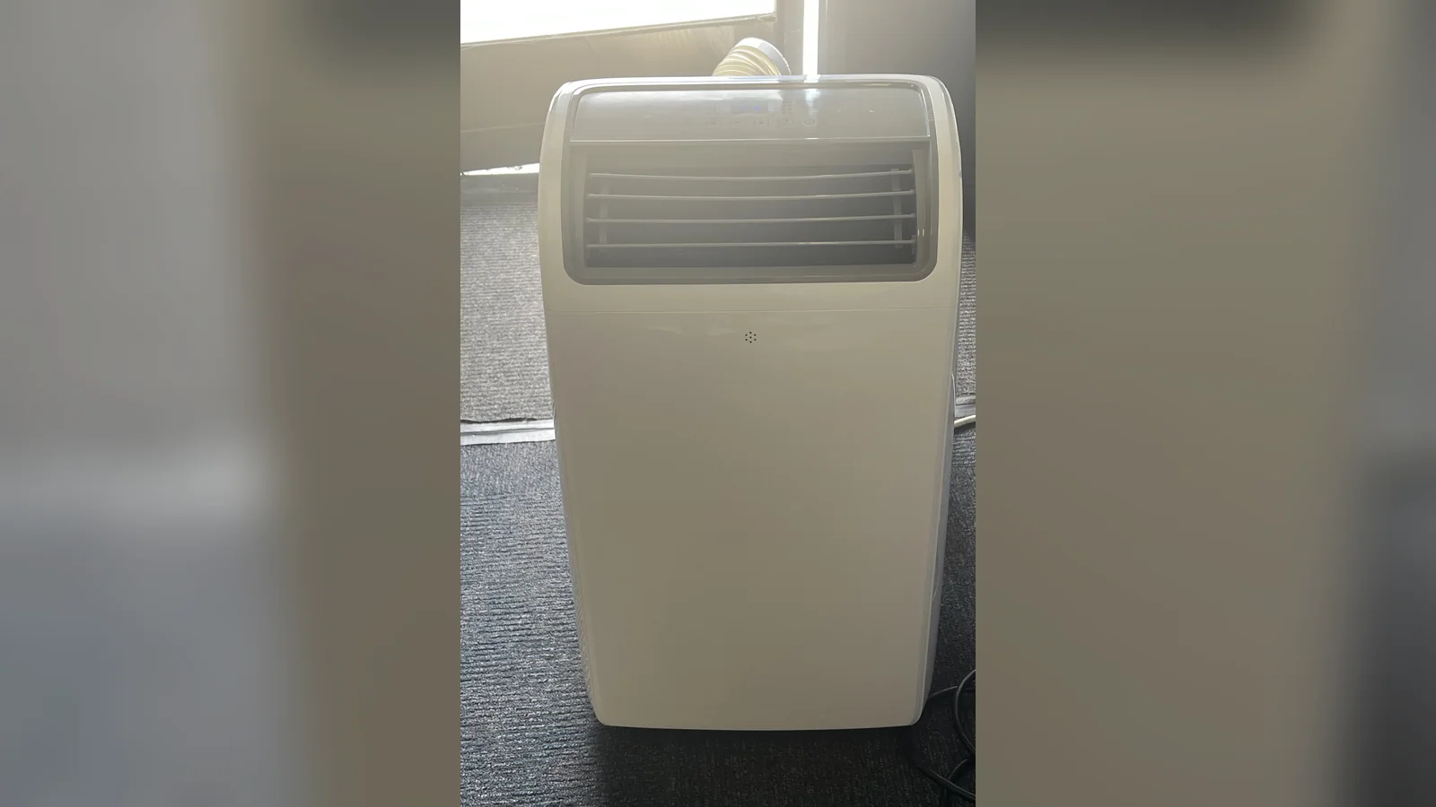 A white portable air conditioner with grey front louvres standing on dark grey carpet, its exhaust hose just visible behind it.