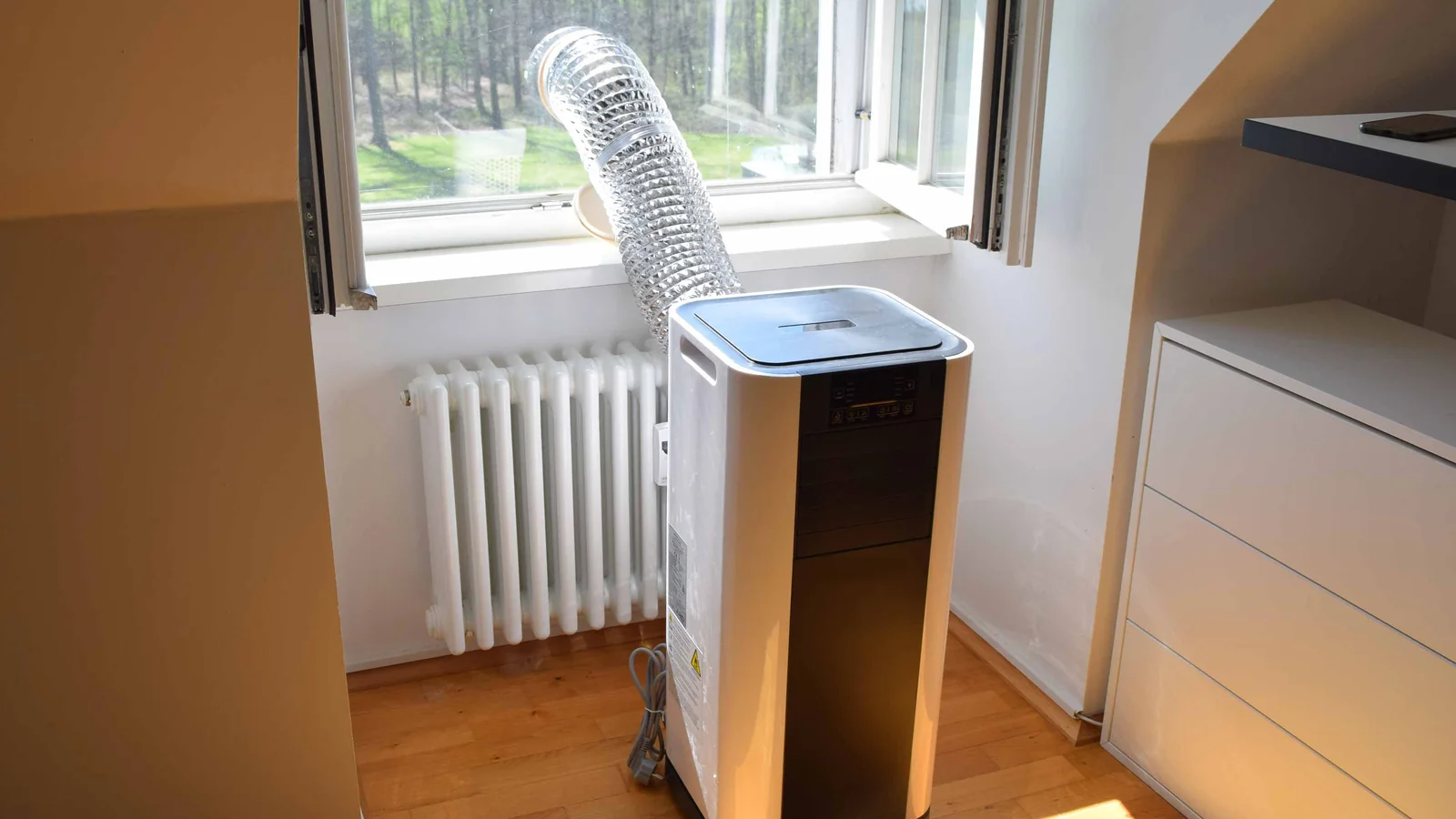 Aircond mudah alih putih dan hitam di atas lantai kayu, hos ekzos perak keluar melalui tingkap yang terbuka.
