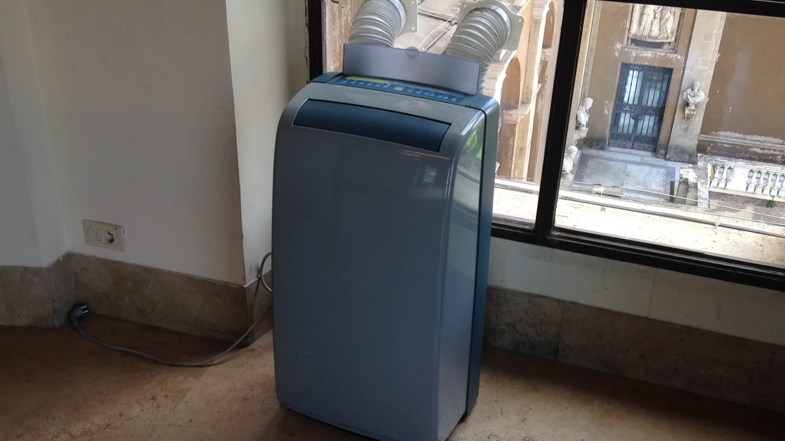 A blue-grey portable air conditioner beside a window, with two exhaust hoses rising from its top.