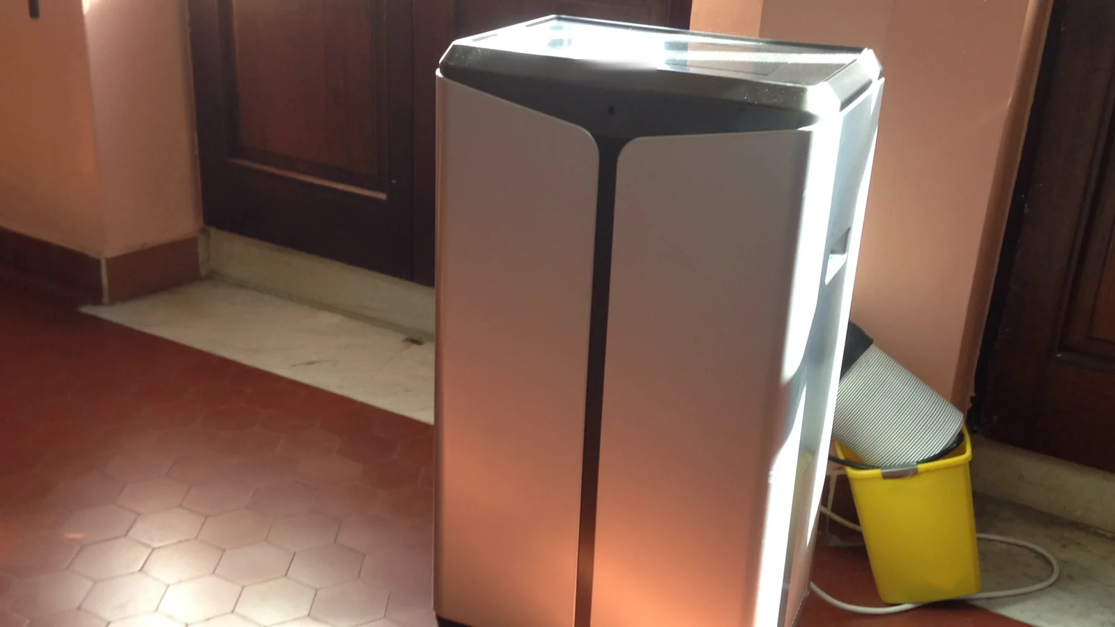 A tall silver portable air conditioner on a tiled floor beside a wooden door, with a bucket behind it.