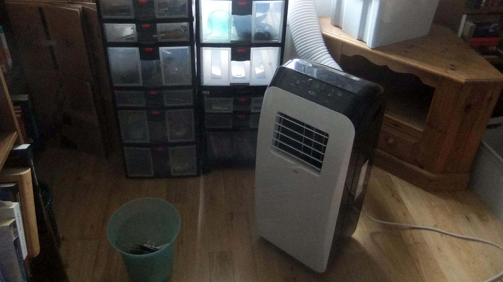 A portable aircon for a small room, with its exhaust hose, standing between plastic storage drawers and a wooden cabinet.