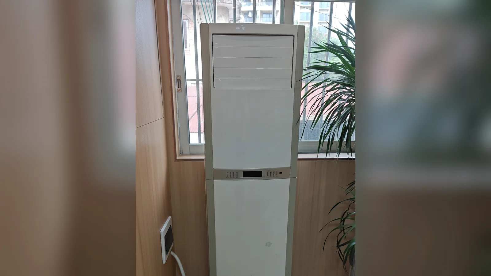 A tall white floor-standing air conditioner in front of a window, next to a potted palm.