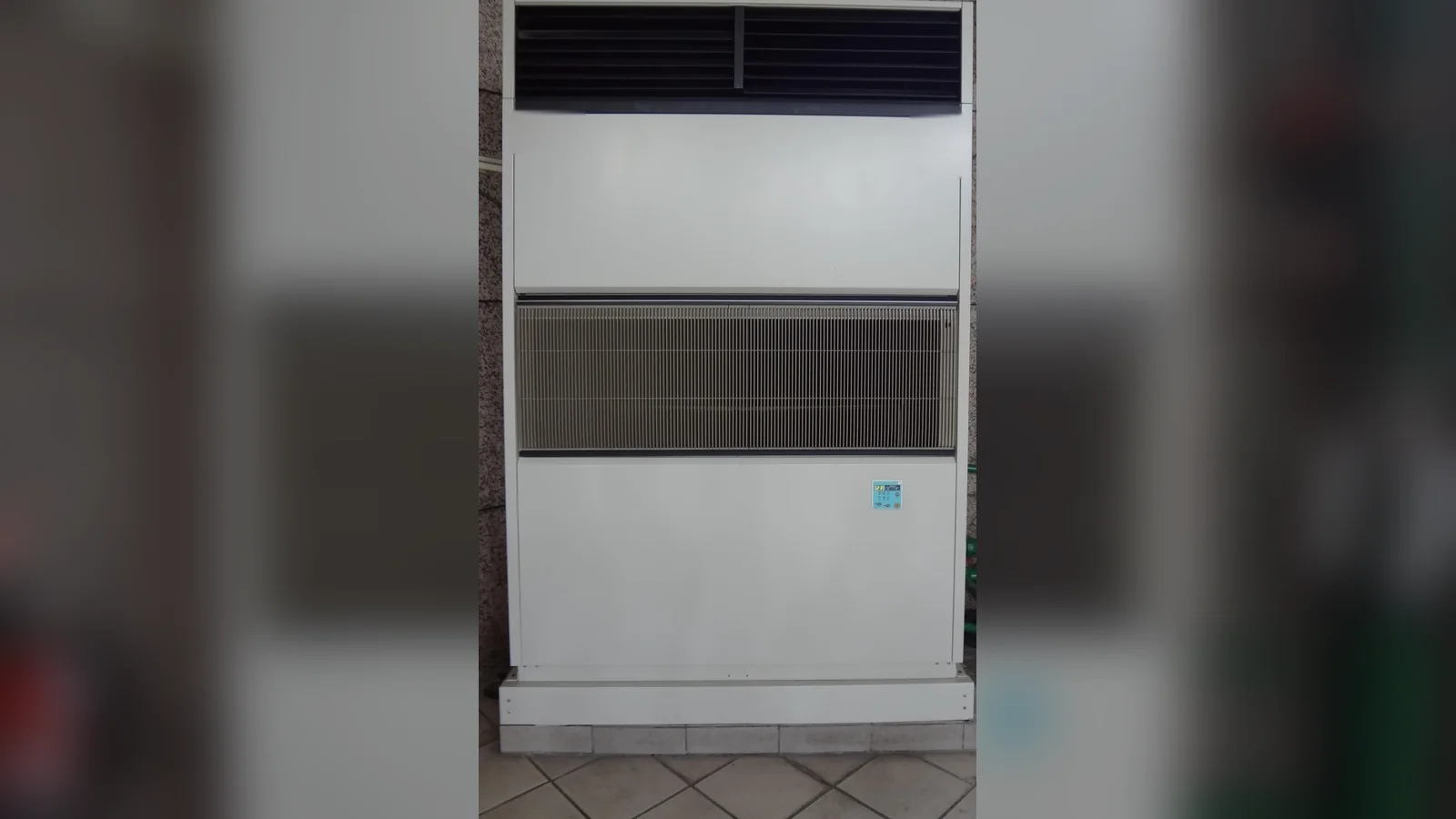 A large floor-standing air conditioner against a granite wall, with a wired controller on its lower panel.