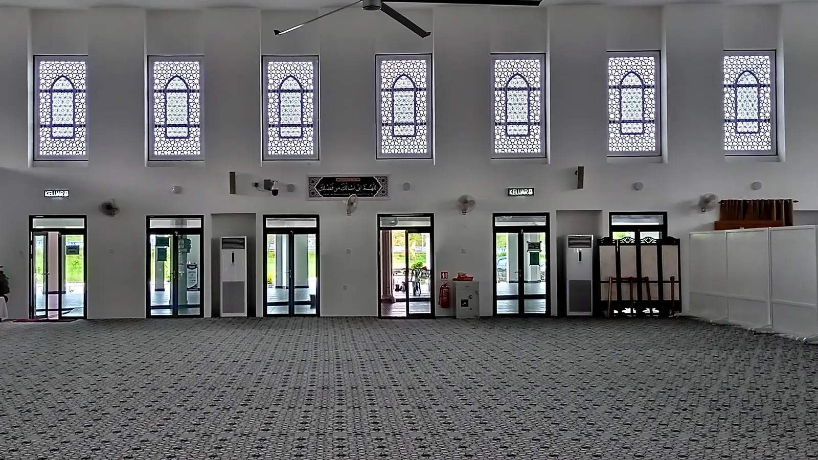 Dewan solat masjid di Malaysia yang kosong, dengan dua aircond masjid jenis berdiri berwarna putih di antara pintu kaca di dinding belakang.