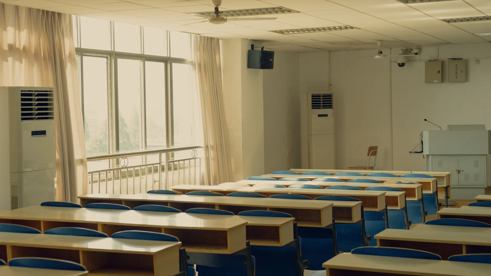Dewan kuliah kosong dengan barisan meja dan dua aircond berdiri yang tinggi di tepi dinding.